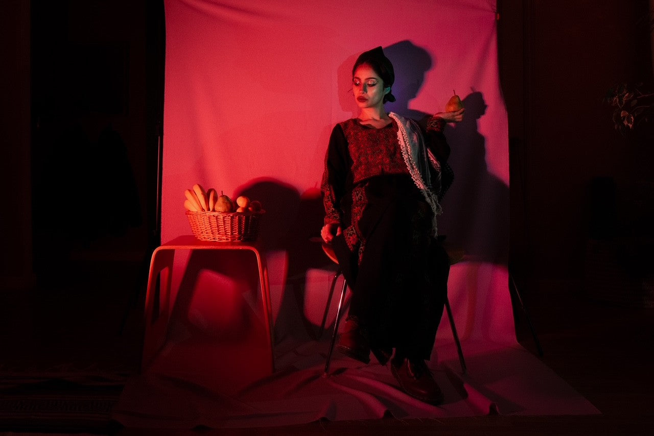 A woman in traditional Palestinian dress sits beneath moody red lighting, holding a pear beside a basket of fruit—evoking themes of heritage, femininity, and still life symbolism.